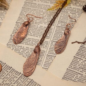 May include: A pair of copper earrings shaped like maple leaves. The earrings are hanging from a book with a dictionary page visible.