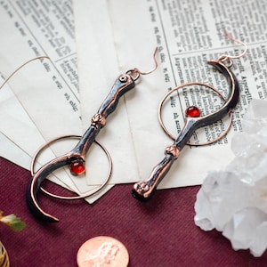 Peut inclure: Une paire de boucles d'oreilles artisanales au design unique. Chaque boucle d'oreille présente un élément incurvé en métal foncé avec des accents cuivrés et une petite pierre précieuse rouge. Les boucles sont suspendues à un cercle cuivré. Une pièce de monnaie est à côté.