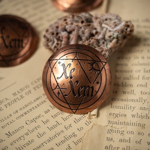 May include: Two copper-colored round metal charms with a pentagram design and the text "Xe Xem" inscribed on them. The charms are on a vintage book page with text.
