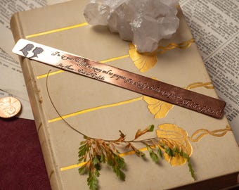 May include: A copper bookmark with a silhouette design and engraved text rests on a vintage book with gold accents. A clear quartz crystal and a penny are also present, along with aged paper in the background. The bookmark features a quote.