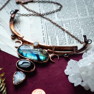 Op de afbeelding: Een handgemaakte koperen ketting met een grote, iriserende labradorietsteen en kleinere zwarte en witte edelstenen. De ketting wordt gepresenteerd op een bordeauxrode ondergrond met een takje groen en een bergkristal.