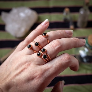May include: A hand wearing five copper rings with black gemstones. The rings are stacked on each other and are worn on the middle finger.