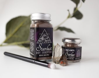 May include: Two glass jars of brown powder labeled "Stardust Dry Shampoo" by Bast & Baetyl Alchemy. The larger jar is 1.0 oz (50g) and the smaller jar is 0.4 oz (12g). A clear quartz crystal is next to the jars.