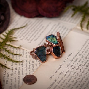 May include: Three copper rings with iridescent green gemstones set in the center of each ring. The rings are arranged on a page of a book with text in Spanish.