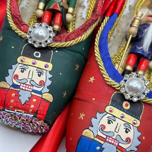 May include: A pair of decorative ballet slippers, one green and one red, each adorned with a hand-painted nutcracker face and a miniature nutcracker figure. The slippers feature gold trim, jewel accents, and red satin ribbons.