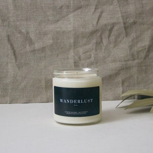 May include: A white candle in a clear glass jar with a black label that reads "WANDERLUST 10 oz.  CHARMING NATURAL SOY CANDLES shopknommeganatural.etsy.com".