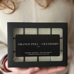 May include: A black box with a clear window showing six white wax melts. The box has a label that reads "Orange Peel + Cranberry 4 oz. Chasing Natural | Soy Wax Melts shopchasingnatural.etsy.com"