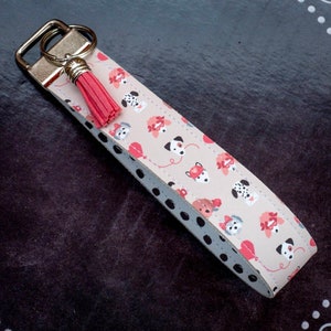 May include: A beige key chain with a pink tassel and a pattern of black and white dogs wearing red bows and holding balloons. The key chain has black polka dots on the edges.
