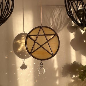 May include: A circular stained glass suncatcher featuring a central five-pointed star design encased in a dark metal frame. The glass is a pale yellow hue with subtle texture. A clear, faceted crystal bead hangs from the bottom of the circle.