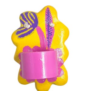 May include: A yellow ceramic wall sconce with a pink painted flower design and a pink painted cylindrical holder. The sconce has a whimsical, abstract shape and is decorated with purple and pink painted leaves.