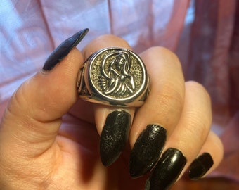 May include: A silver ring with a detailed design of a woman holding a scythe. The ring is on a finger with black nail polish.