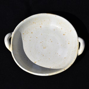 Peut inclure: Un plat à four en céramique blanche avec deux poignées. Le plat a un aspect rustique et moucheté avec des taches brunes.