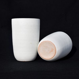 Peut inclure: Deux tasses en céramique blanche avec une finition lisse et mate. La tasse de droite a une base en argile brune avec une marque de fabricant.