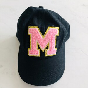Puede incluir: Gorra de b&eacute;isbol negra con una letra M rosa y dorada en la parte delantera.