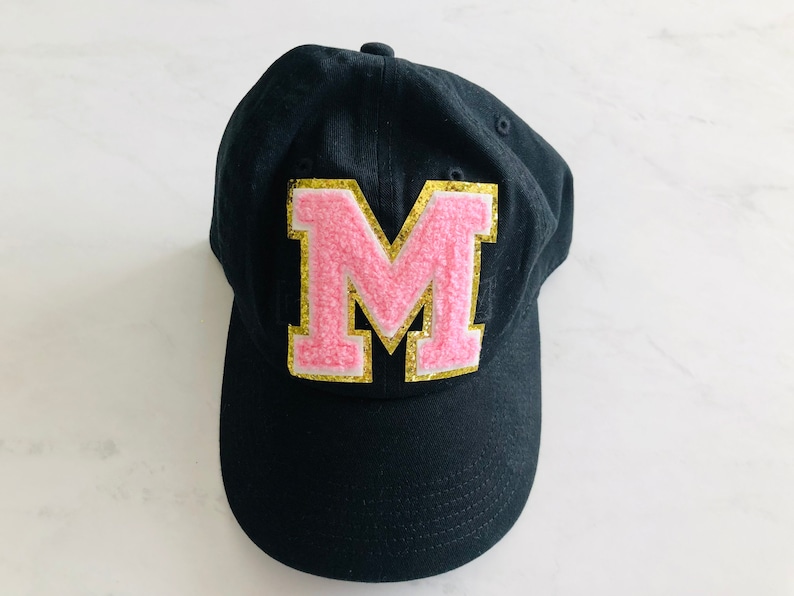 Puede incluir: Gorra de b&eacute;isbol negra con una letra M rosa y dorada en la parte delantera.