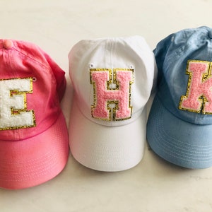Puede incluir: Tres gorras de b&eacute;isbol de tela rosa, blanca y azul. Cada gorra tiene una gran letra rosa con ribete dorado de purpurina en la parte delantera. Las letras son "E", "H" y "K".