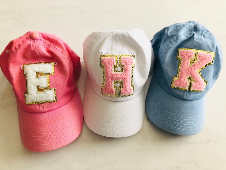 Puede incluir: Tres gorras de b&eacute;isbol de tela rosa, blanca y azul. Cada gorra tiene una gran letra rosa con ribete dorado de purpurina en la parte delantera. Las letras son "E", "H" y "K".