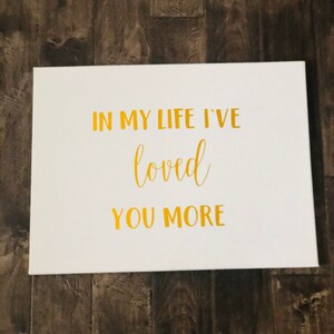May include: White canvas with gold lettering that reads "In my life I've loved you more".