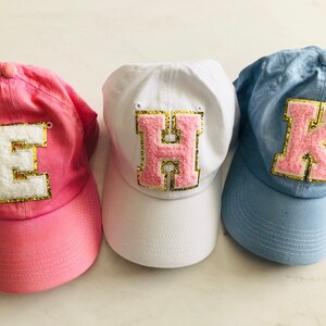 Puede incluir: Tres gorras de b&eacute;isbol de tela rosa, blanca y azul. Cada gorra tiene una letra grande, rosa y peluda cosida en la parte delantera. Las letras son E, H y K.