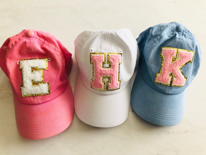 Puede incluir: Tres gorras de b&eacute;isbol de tela rosa, blanca y azul. Cada gorra tiene una letra grande, rosa y peluda cosida en la parte delantera. Las letras son E, H y K.