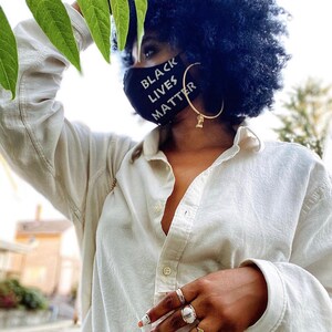 May include: A person wearing a white button-down shirt with a black face mask that says "Black Lives Matter". The person is wearing gold hoop earrings and several rings on their fingers.