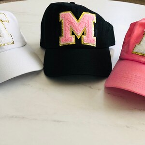 Puede incluir: Cinco gorras de b&eacute;isbol con diferentes letras en la parte delantera. Las letras son K, T, M, E y H. Las letras son de color rosa con ribetes dorados. Las gorras son de diferentes colores: azul, blanco, negro, rosa y blanco.