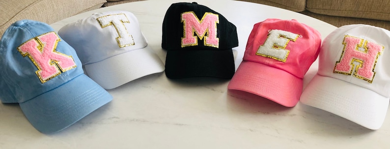 Puede incluir: Cinco gorras de b&eacute;isbol con diferentes letras en la parte delantera. Las letras son K, T, M, E y H. Las letras son de color rosa con ribetes dorados. Las gorras son de diferentes colores: azul, blanco, negro, rosa y blanco.