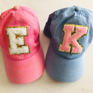 Puede incluir: Dos gorras de b&eacute;isbol, una rosa con una "E" blanca y una azul con una "K" rosa, ambas con ribete de purpurina dorada.