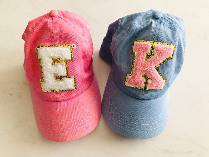 Puede incluir: Dos gorras de b&eacute;isbol, una rosa con una "E" blanca y una azul con una "K" rosa, ambas con ribete de purpurina dorada.