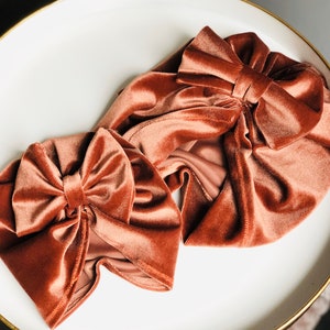 Puede incluir: Dos diademas de terciopelo de color naranja quemado. Ambas diademas tienen un gran lazo en la parte superior. Las diademas est&aacute;n sobre una superficie blanca.