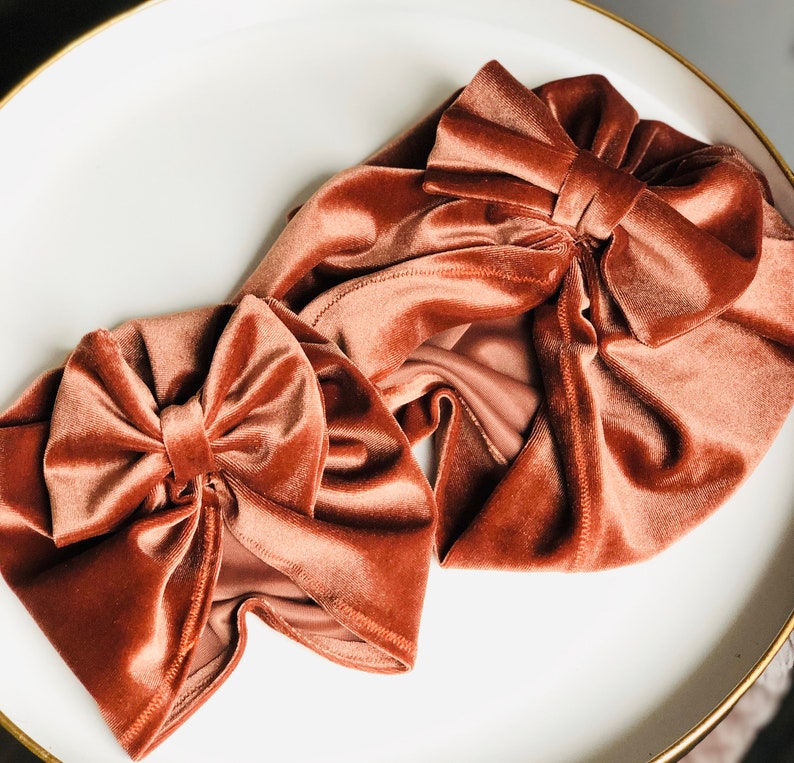 Puede incluir: Dos diademas de terciopelo de color naranja quemado. Ambas diademas tienen un gran lazo en la parte superior. Las diademas est&aacute;n sobre una superficie blanca.