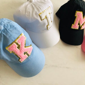 Puede incluir: Tres gorras de b&eacute;isbol con diferentes colores de tela y letras rosas y doradas. Las gorras son blancas, azul claro y negras. Las letras son K, M y T.