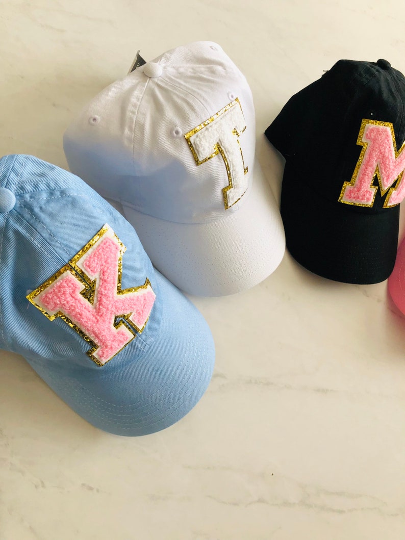 Puede incluir: Tres gorras de b&eacute;isbol con diferentes colores de tela y letras rosas y doradas. Las gorras son blancas, azul claro y negras. Las letras son K, M y T.
