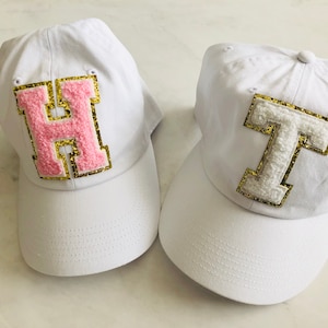 Puede incluir: Dos gorras de b&eacute;isbol blancas con letras bordadas. La primera gorra tiene una "H" rosa con ribete dorado. La segunda gorra tiene una "T" blanca con ribete dorado.