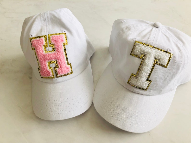 Puede incluir: Dos gorras de b&eacute;isbol blancas con letras bordadas. La primera gorra tiene una "H" rosa con ribete dorado. La segunda gorra tiene una "T" blanca con ribete dorado.