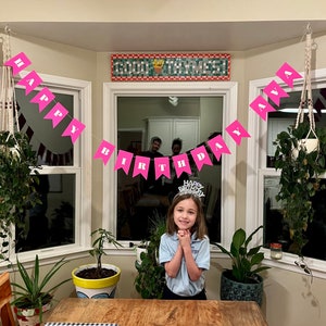 May include: A young girl wearing a blue shirt and a birthday crown stands in front of a table with a pink banner that reads "Happy Birthday Ava".