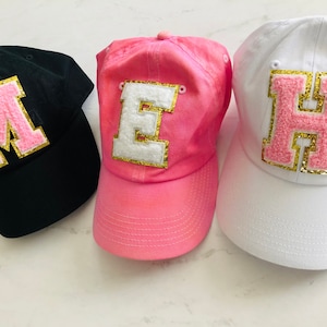 Puede incluir: Tres gorras de b&eacute;isbol con grandes letras de fieltro en la parte delantera. Las gorras son negras, rosas y blancas. Las letras son rosas con ribetes dorados. Las letras deletrean "MEH".