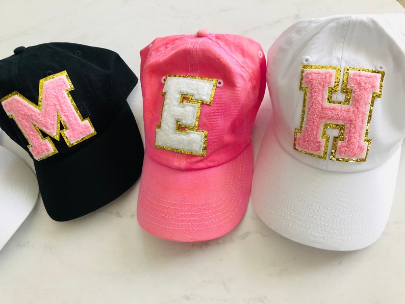 Puede incluir: Tres gorras de b&eacute;isbol con grandes letras de fieltro en la parte delantera. Las gorras son negras, rosas y blancas. Las letras son rosas con ribetes dorados. Las letras deletrean "MEH".
