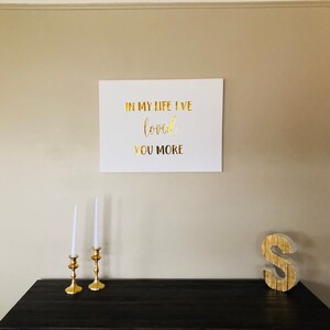 May include: A white canvas with gold lettering that reads "In my life I've loved you more". The canvas is hanging on a beige wall above a dark wood table with two gold candle holders and a gold letter S.