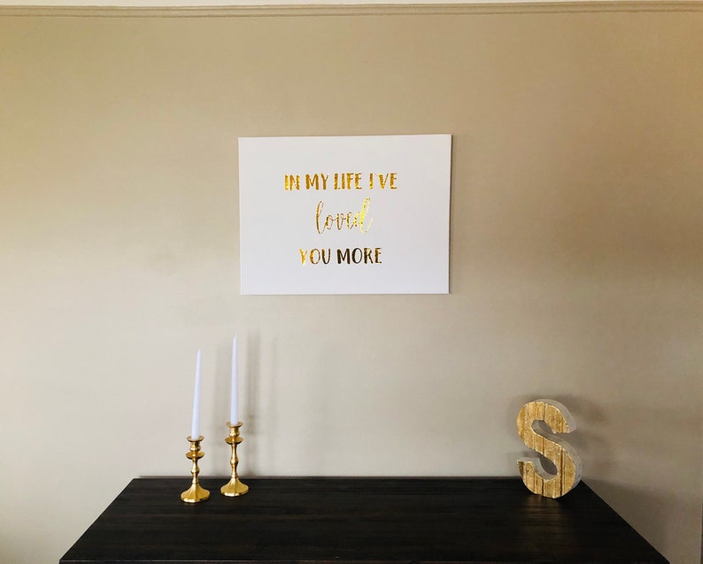 May include: A white canvas with gold lettering that reads "In my life I've loved you more". The canvas is hanging on a beige wall above a dark wood table with two gold candle holders and a gold letter S.
