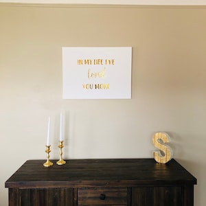 May include: A white canvas with gold lettering that reads "In my life I've loved you more". The canvas is hanging on a beige wall above a dark brown wooden dresser with three drawers. Two gold candle holders with white candles are on the dresser. A gold letter "S" is on the dresser to the right of the candle holders.