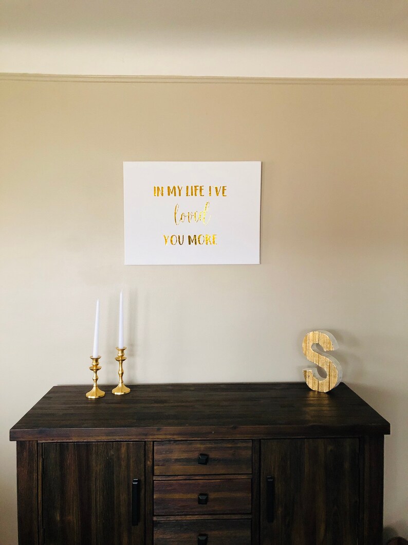 May include: A white canvas with gold lettering that reads "In my life I've loved you more". The canvas is hanging on a beige wall above a dark brown wooden dresser with three drawers. Two gold candle holders with white candles are on the dresser. A gold letter "S" is on the dresser to the right of the candle holders.