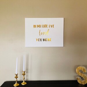 May include: A white canvas with gold lettering that reads "In my life I've loved you more". The canvas is hanging on a beige wall. There are two gold candle holders with white candles on a black table in the foreground. A gold letter "S" is on the table to the right.