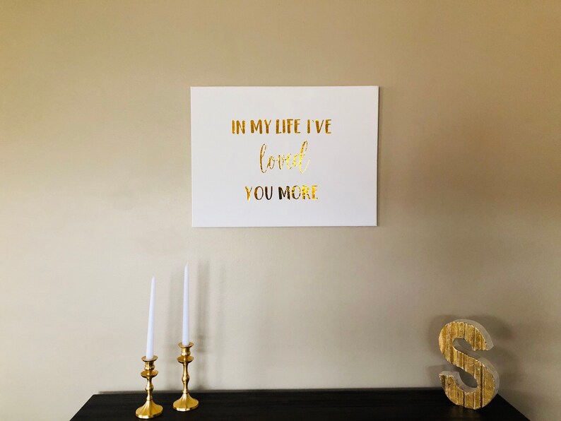 May include: A white canvas with gold lettering that reads "In my life I've loved you more". The canvas is hanging on a beige wall. There are two gold candle holders with white candles on a black table in the foreground. A gold letter "S" is on the table to the right.