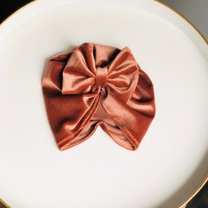 Puede incluir: Un turbante de terciopelo terracota con un gran lazo en la parte superior. El turbante est&aacute; sobre una superficie blanca.