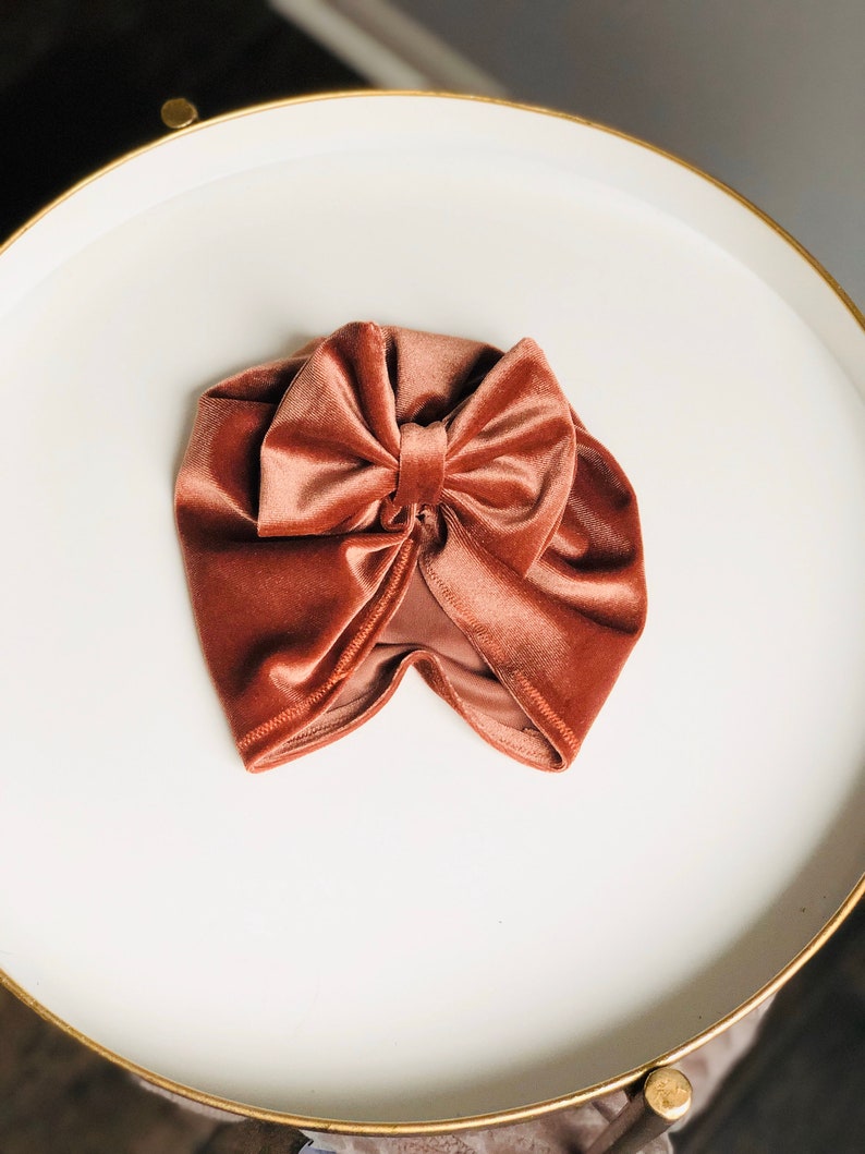 Puede incluir: Un turbante de terciopelo terracota con un gran lazo en la parte superior. El turbante est&aacute; sobre una superficie blanca.