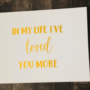 May include: White canvas with gold lettering that reads "In my life I've loved you more".