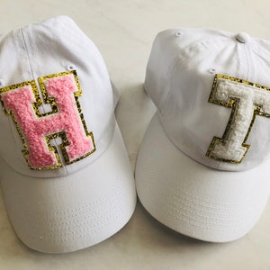 Puede incluir: Dos gorras de b&eacute;isbol blancas con letras bordadas. La gorra de la izquierda tiene una "H" rosa con ribete dorado de purpurina. La gorra de la derecha tiene una "T" blanca con ribete dorado de purpurina.