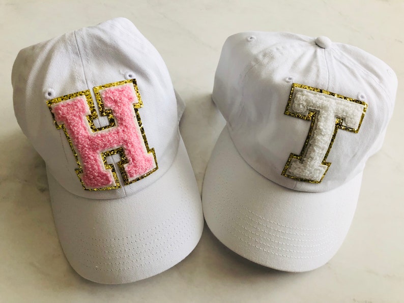Puede incluir: Dos gorras de b&eacute;isbol blancas con letras bordadas. La gorra de la izquierda tiene una "H" rosa con ribete dorado de purpurina. La gorra de la derecha tiene una "T" blanca con ribete dorado de purpurina.