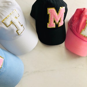 Puede incluir: Cuatro gorras de b&eacute;isbol con diferentes colores de tela y grandes letras de fieltro en la parte delantera. Las gorras son blancas, negras, rosas y azul claro. Las letras son T, M, L y J. Las letras est&aacute;n bordeadas con purpurina dorada.